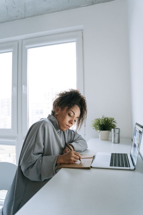 Woman in casual clothing organizing notes beside a laptop in a bright workspace.