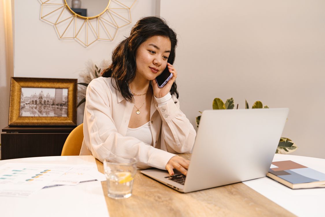 Woman in casual clothing talking on a mobile phone while working on a laptop.